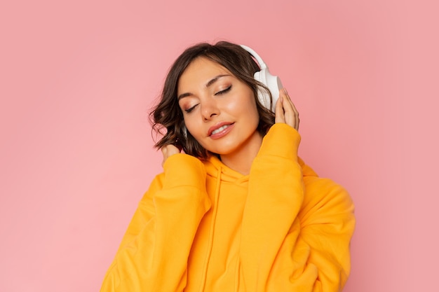Blissful smiling woman in white earphones listening to music on pink. Wearing orange hoodie.