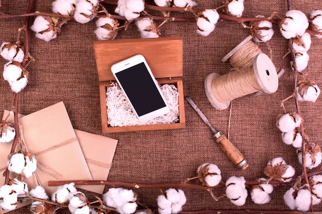 Bloem met pluizige gedroogde katoenen bollen geschenkdozen, witte smartphone en jute touw hank over ruwe bruine jute