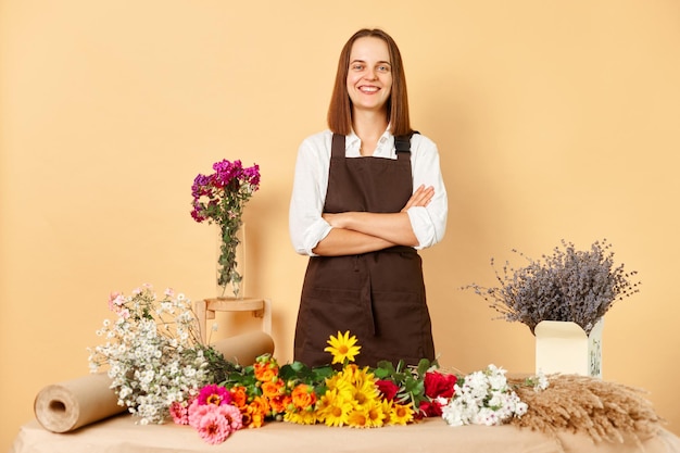 Bloemhandeldiensten Bloemenwinkel ambiance Verse bloemen te koop Zeker glimlachende bruinharige vrouw bloemhandelaar die op haar werkplaats een bruine schort draagt en tegen een beige muur poseert