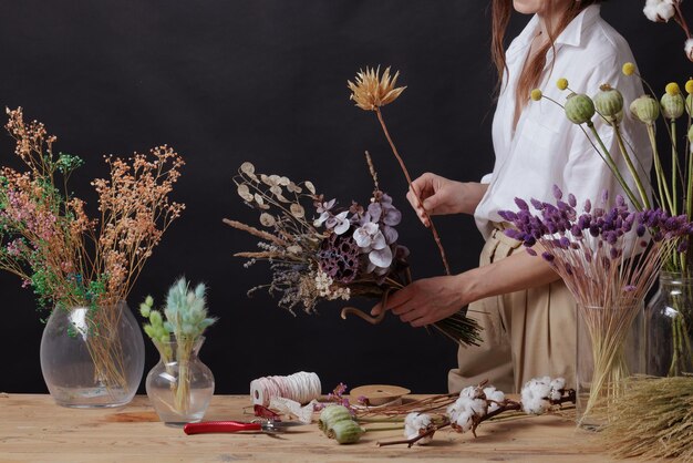 Bloemist maakt een boeket gedroogde bloemen aan een houten tafel op een effen donkere achtergrond met copyspace
