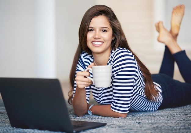 Bloggen met een kopje koffie Portret van een lachende jonge vrouw die thuis op de vloer ligt met een laptop