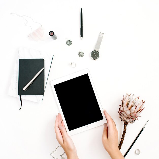 Blogger or freelancer workspace with blank screen tablet, protea flower, notebook, watches and feminine accessories on white background. Flat lay, top view minimalistic decorated home office desk.