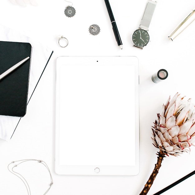 Blogger or freelancer workspace with blank screen tablet, protea flower, notebook, watches and feminine accessories on white background. Flat lay, top view minimalistic decorated home office desk.