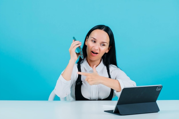 Blogger girl is holding phone and pointing left with forefinger by sitting in front of tablet camera on blue background