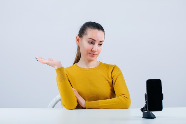 Blogger girl is pointing left with hand by sitting in front of mobile camera on white background