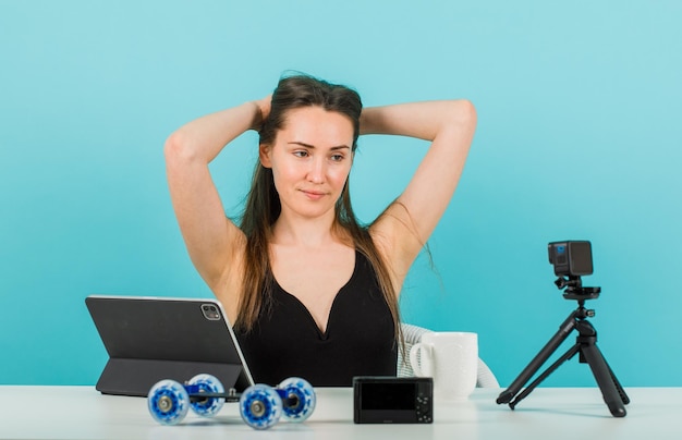 Blogger girl is posing at camera by holding hands behing head on blue background