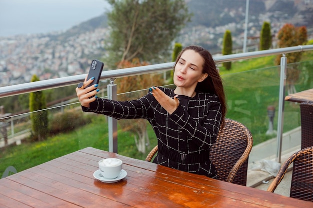 ブロガーの女の子は、街の景色の背景に座ってキスを吹いてスマートフォンで自分撮りをしています