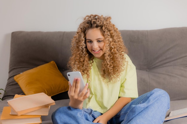 Blogger girl with headphones and a smartphone in her hands is sitting cross-legged on a gray sofa. young woman with curly hair communicates online. High quality photo