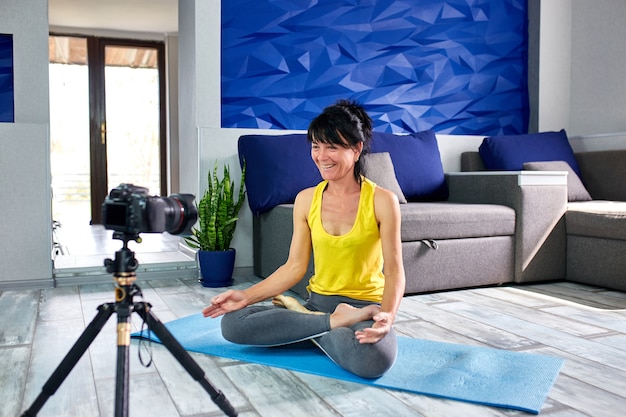 Blogger senior woman with slim body shape in sportswear doing yoga at home. Fitness blog, video tutorial, online training recording a video yoga lesson by camera.