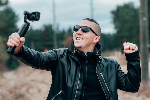 Blogger in occhiali da sole che fa selfie o video in streaming nella foresta di pini utilizzando la action cam con stabilizzatore della fotocamera cardanica.