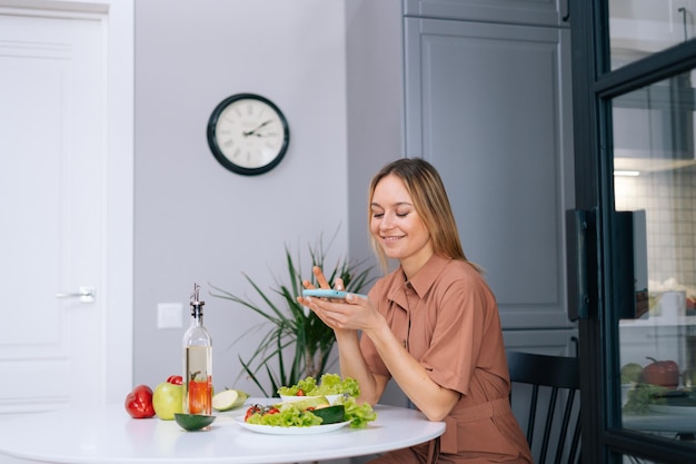 Фото Молодая женщина-блогер использует смартфон, чтобы фотографировать овощи, лежащие на кухонном столе. концепция социальных сетей. концепция здорового питания.