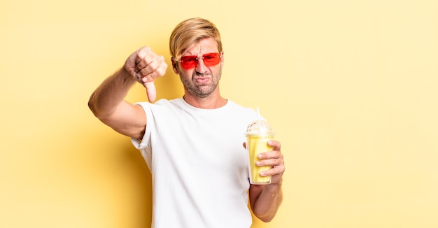 Blond adult man feeling cross,showing thumbs down with a milkshake