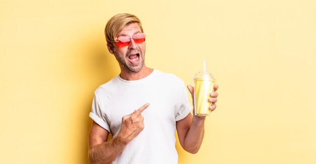 Blond adult man looking excited and surprised pointing to the side with a milkshake