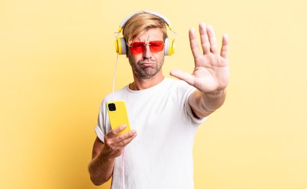 Blond adult man looking serious showing open palm making stop gesture with headphones