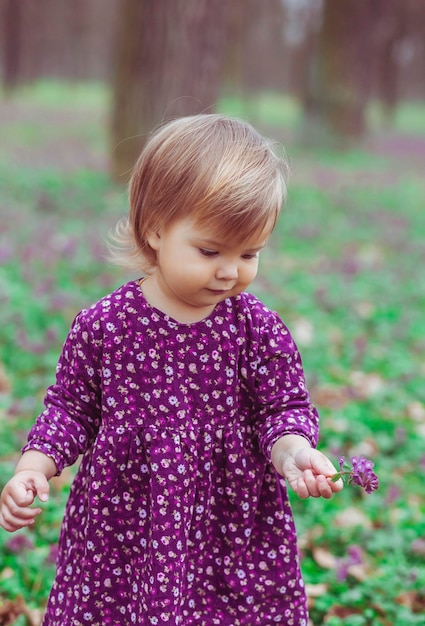 blond baby in a colored dress holding flower in a forest glade