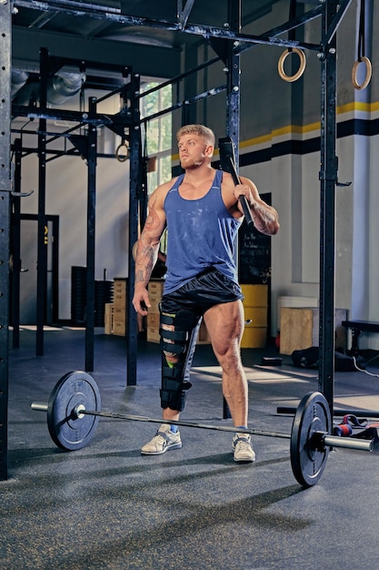 Blond bodybuilder with broken leg in bandage holds cross fit hammer in a gym club.