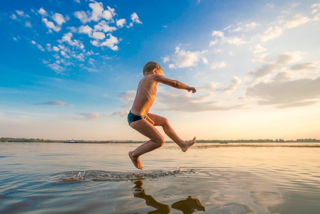 Blond boy in a black bathing jumping into the water against the backdrop of blue sky with clouds in the sunlight