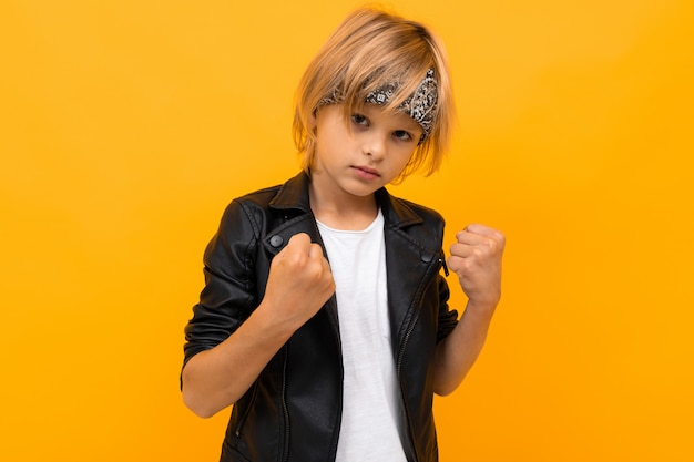 Photo blond boy in a black leather jacket and a white t-shirt with a bandana holds his fists on orange
