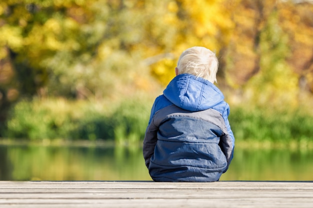 Photo blond boy in blue jacket sitting on the river pier. back view