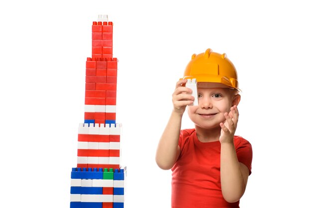 Photo blond boy in construction helmet and red t-shirt builds a skyscraper from the details of the designer. isolate on white background.