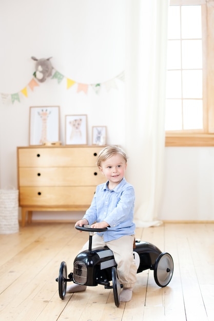 Blond Boy drives a retro toy car in his children room. Cute baby playing in kindergarten. Childhood concept. Child development concept. Bright children's room in the Scandinavian style. Decor room