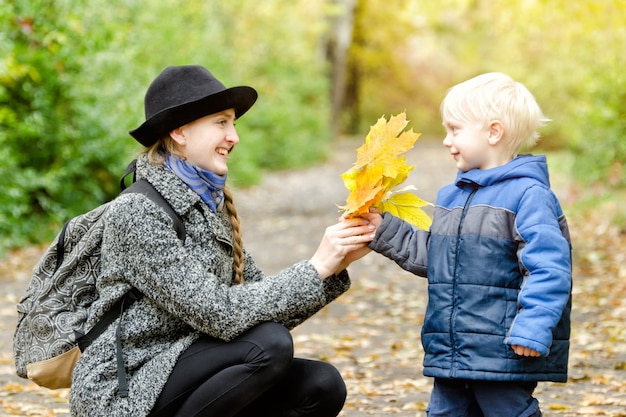 Blond boy gives his mother a bouquet of yellow leaves. Autumn forest