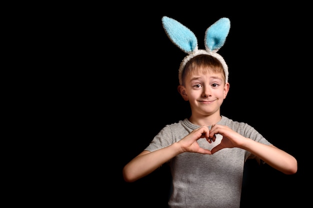 Blond boy in hare ears folded his hands in heart shape on his chest on black