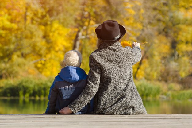 Blond boy and his mother sitting on the dock. Autumn day. Back view