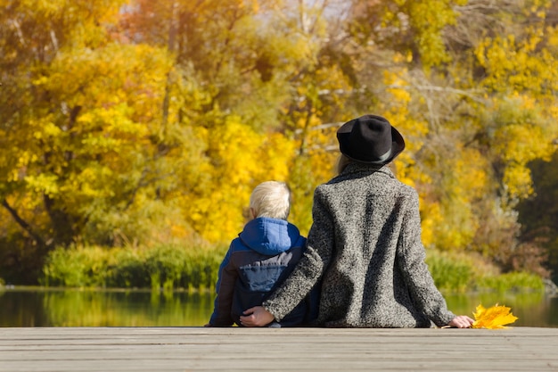 Blond boy and his mother sitting on the dock. Autumn day. Back view