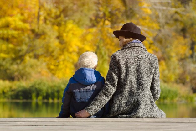 ブロンドの男の子と彼の母親がドックに座っている秋の日 後ろの景色