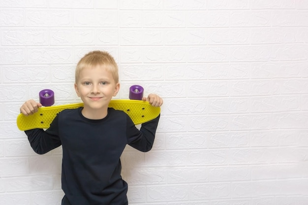 Blond boy holding a skateboard behind his shoulders smiling against the background of a white wall with copyspace teenagers activity