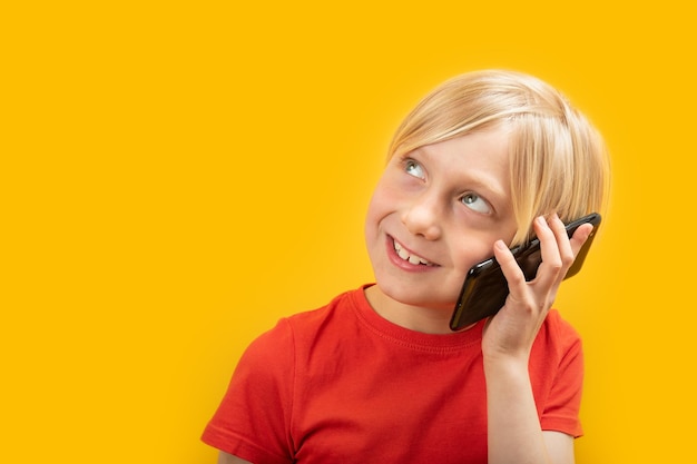 Фото Блондин в красной футболке разговаривает по телефону, улыбаясь и глядя вверх портрет ребенка с изоляцией смартфона на желтом фоне скопируйте пространство