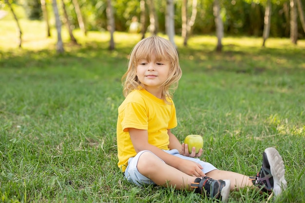 a blond boy is sitting on the lawn in a yellow T-shirt holding a green apple in his hands