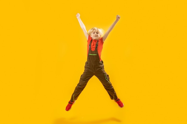 Blond boy jumps high with his hands up Portrait of schoolboy in overalls bouncing on yellow background Copy space