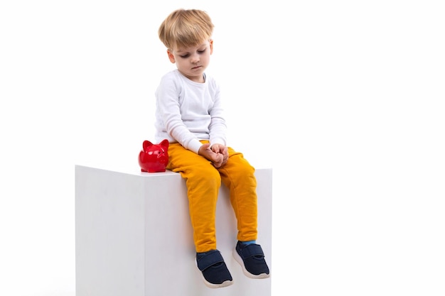 A blond boy sits on a white background next to a red piggy bank and is bored