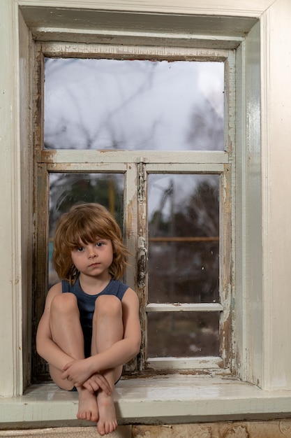 Blond boy sits on a window with a wooden common sill