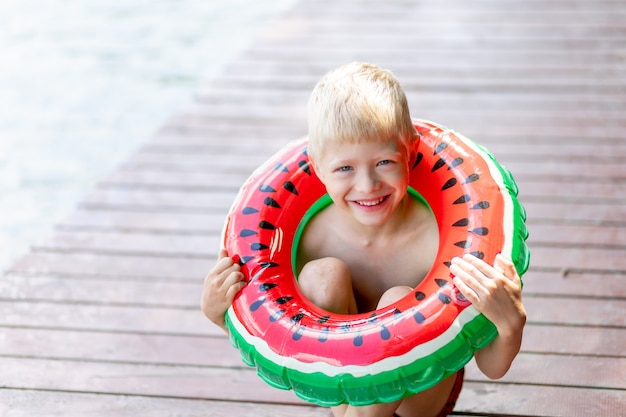 белокурый мальчик сидит на пирсе у воды с красно-зеленым поплавком
