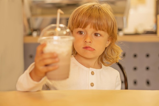 a blond boy in a white linen shirt holds a cafe with a milkshake