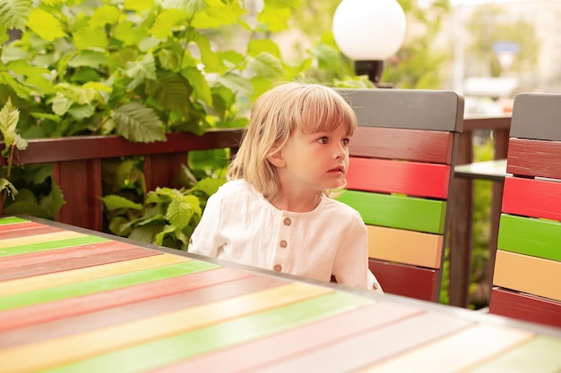 a blond boy in a white linen shirt sits at a table in an outdoor cafe and looks at the table