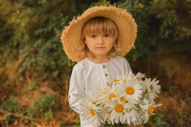 a blond boy in a white linen suit and a straw hat holds a bouquet of daisies in his hands