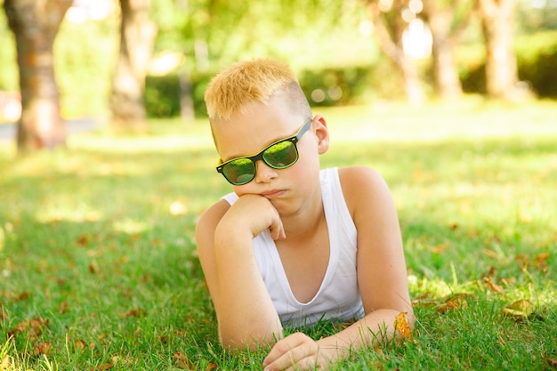 A blond boy in a white T-shirt with sunglasses lies on the grass in the park