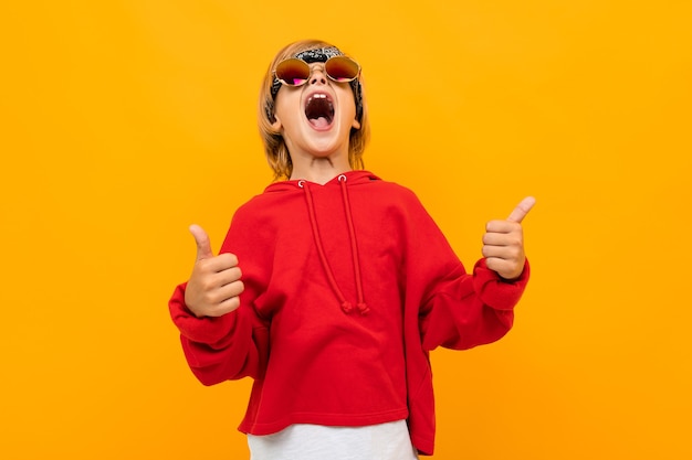 Blond boy with a bandana on his head in a red sweater and glasses shows a class on an orange background.