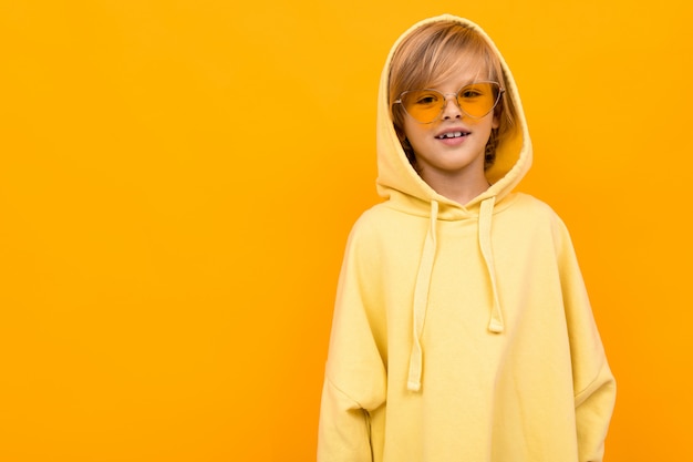 Blond boy with a panama in a light hoodie with glasses posing on yellow studio