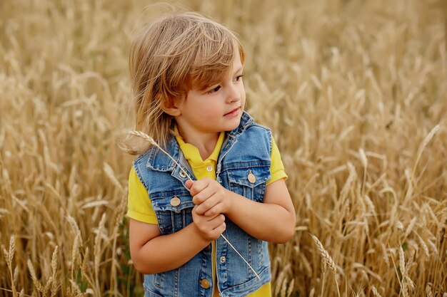 a blond boy in a yellow jacket a denim vest and linen shorts is standing by a haystack