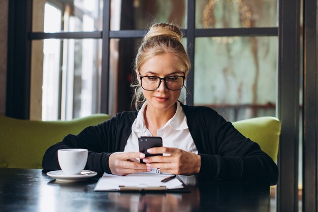 Blond business woman sitting on workspace and using laptop at office, she looks at a folder with documents, talking by phone and drinking cup of coffee.