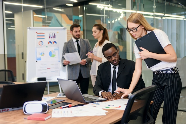 Blond businesswoman in stylish glasses gives for signature different documents to purposeful dark-skinned manager in the meeting room with other team mates at the background.