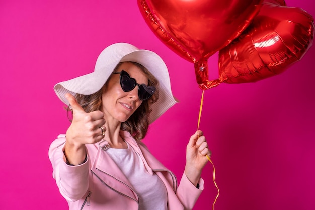 Blond caucasian woman in a white hat and sunglasses making the ok sign with her hands in a nightclub with some heart balloons taking a selfie pink background