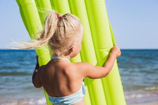 Blond child girl in a swimsuit holding an inflatable mattress on the beach.