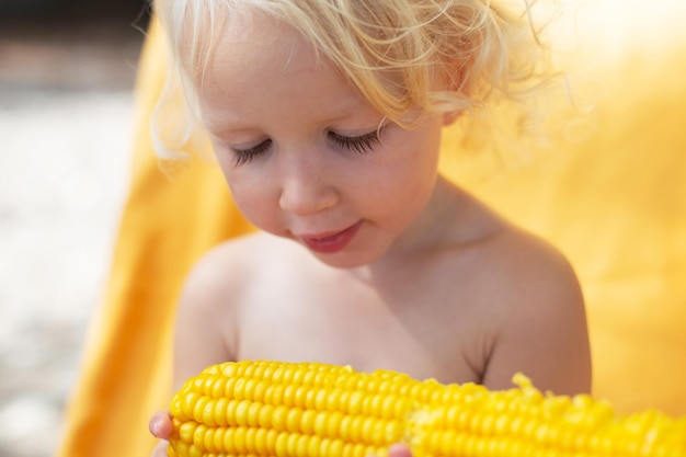 Blond and curly toddler girl eating corn on the beach yellow colors summer mood