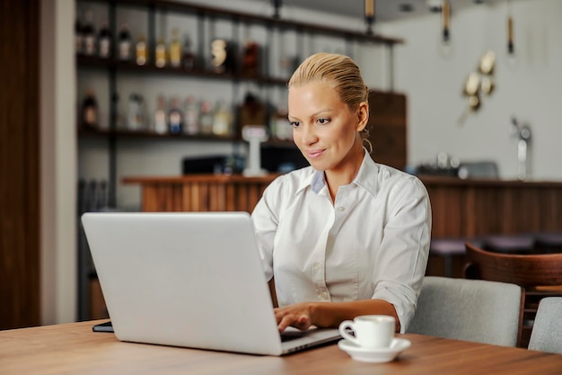 A blond elegant businesswoman is sitting in working friendly cafe and typing a report on her laptop